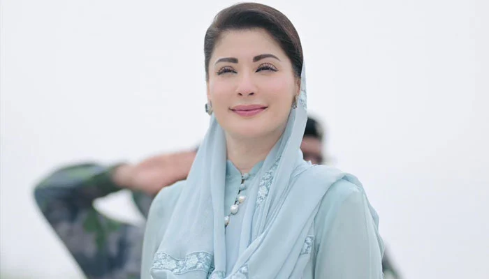 A woman in a light blue outfit and scarf stands solemnly, with a man in military uniform saluting in the background.
