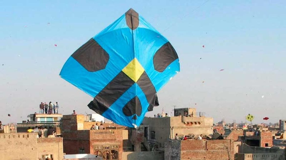 A vibrant blue kite with black and yellow accents soars over rooftops, surrounded by fluttering kites in a clear sky.