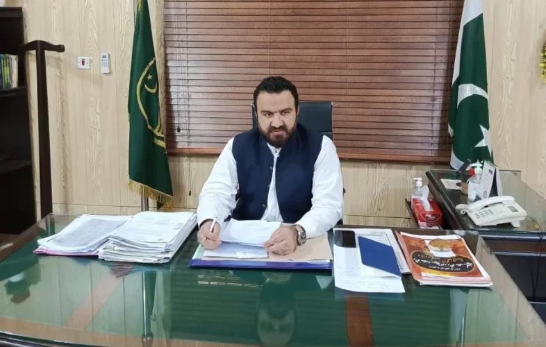 A person in formal attire sits at a desk covered with documents and a telephone, with a green and white flag in the background.