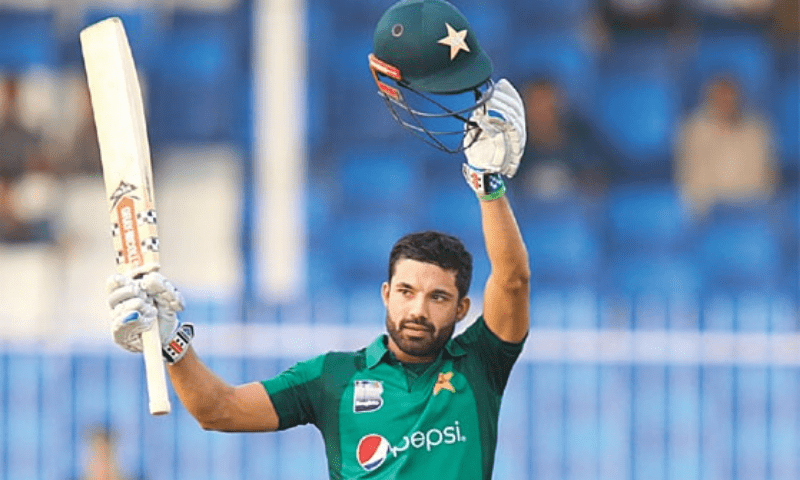 A cricketer in a green jersey celebrates a milestone by raising his bat and helmet on the field, showcasing team pride.