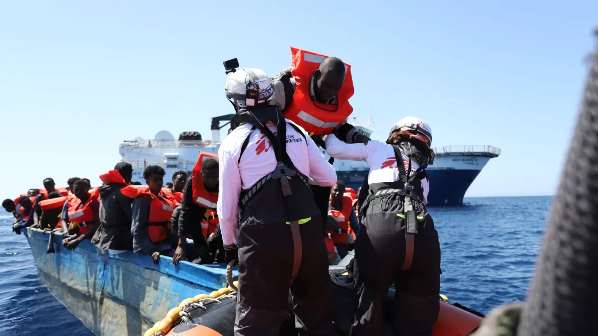 Rescue workers assist migrants in life jackets aboard a small boat, with a larger ship visible in the background on a clear day.