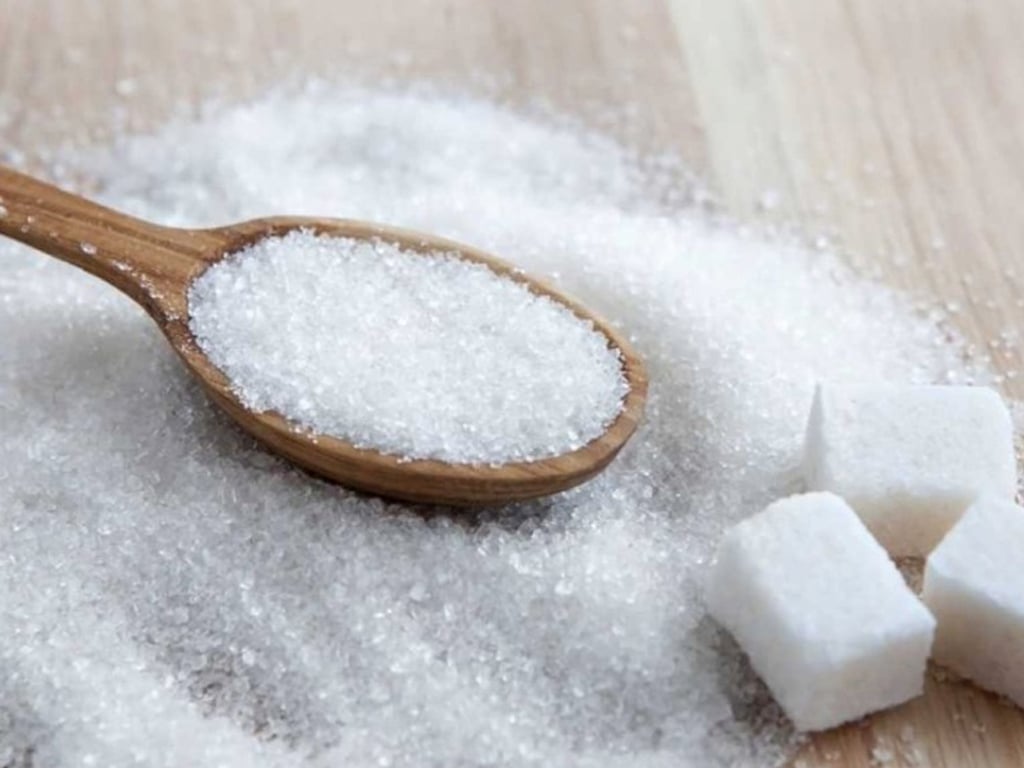 A wooden spoon filled with granulated sugar rests on a surface scattered with sugar and three sugar cubes nearby.