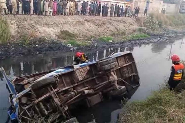 Rescue workers in helmets assist in recovering a flipped-over vehicle from a stagnant, polluted waterway, while onlookers watch from the bank.