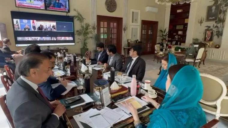 A group of professionals engaged in a meeting, with documents and beverages on a large table, and a video conference displayed on a screen.
