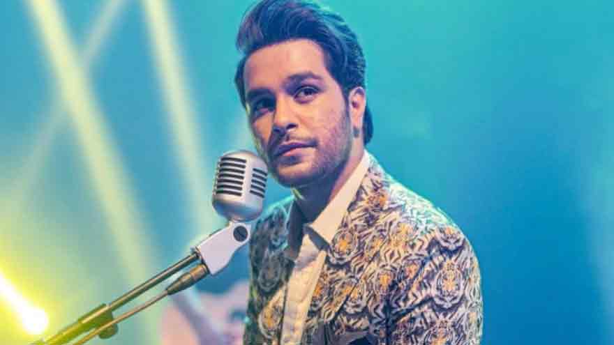 A performer in a patterned jacket sings into a vintage microphone against a vibrant, colorful light backdrop.
