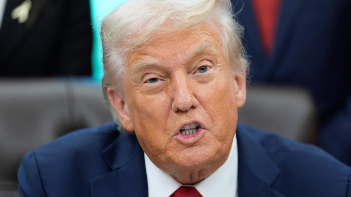 A man in a blue suit with a red tie sits at a table, with a blurred face and other people visible in the background.