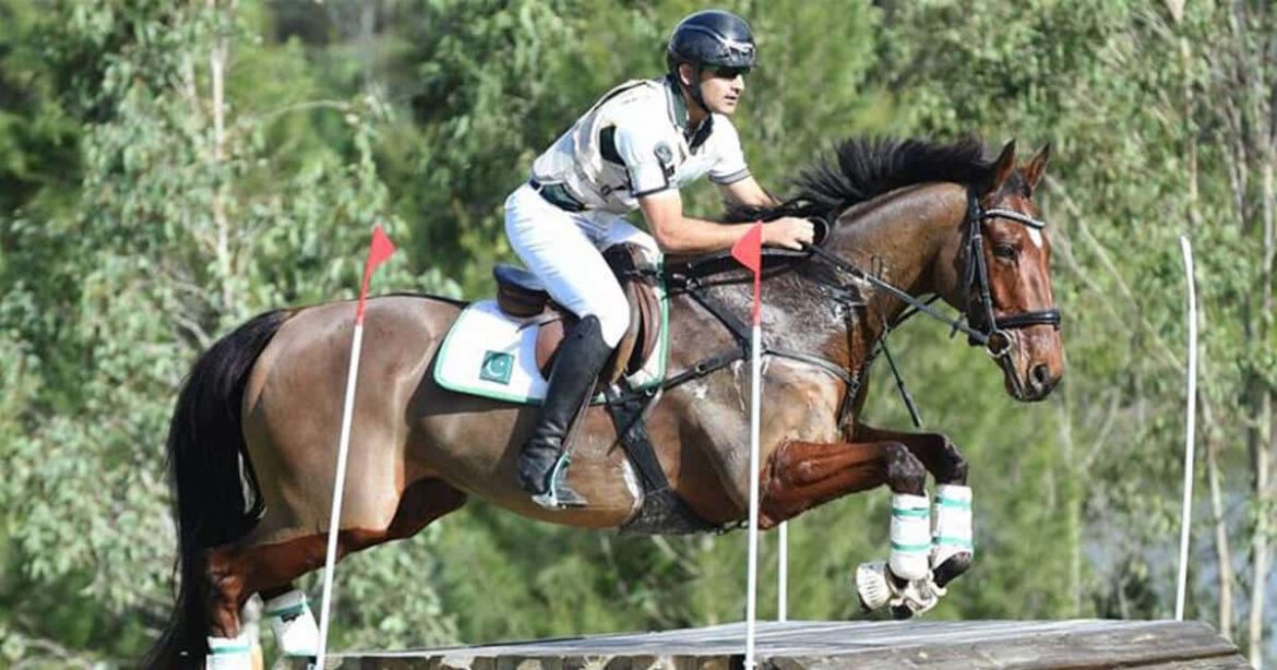 A rider in a green and white uniform guides a jumping horse over a wooden obstacle, flanked by red flags and lush greenery.