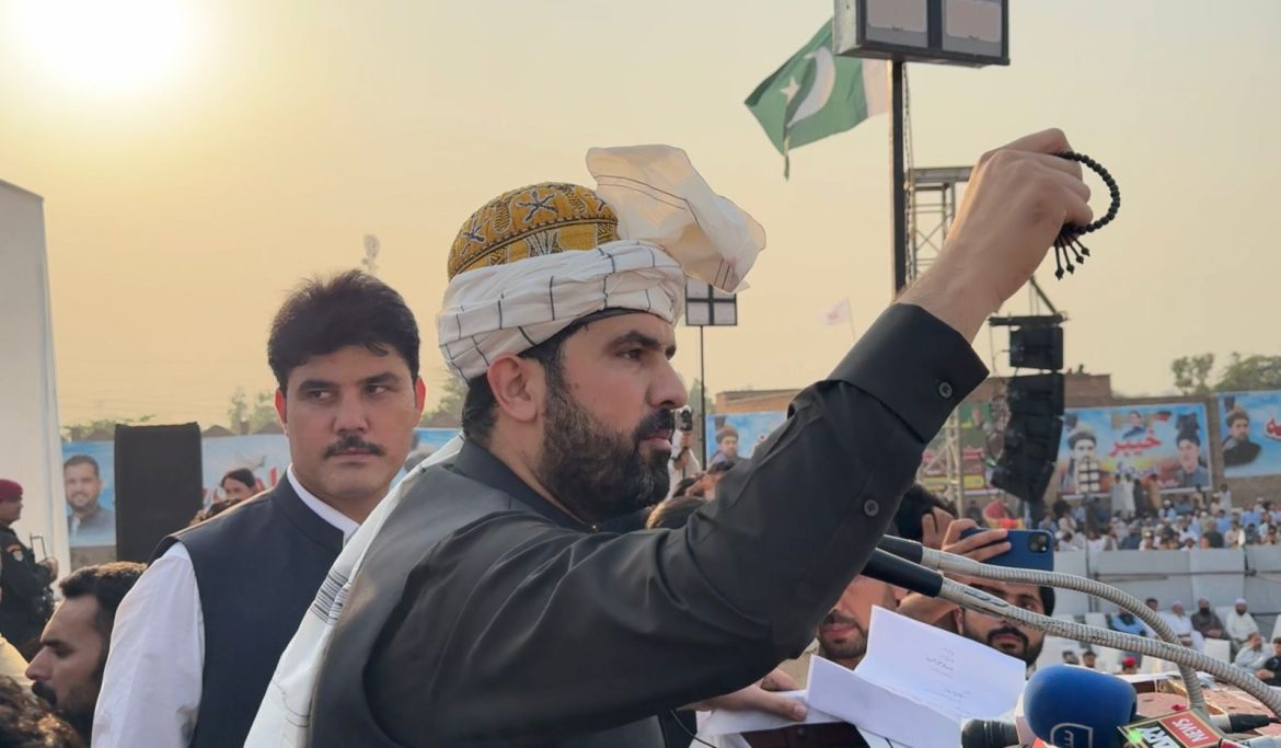 A speaker gestures passionately while holding a prayer bead, with a crowd and flags in the background at an outdoor event.