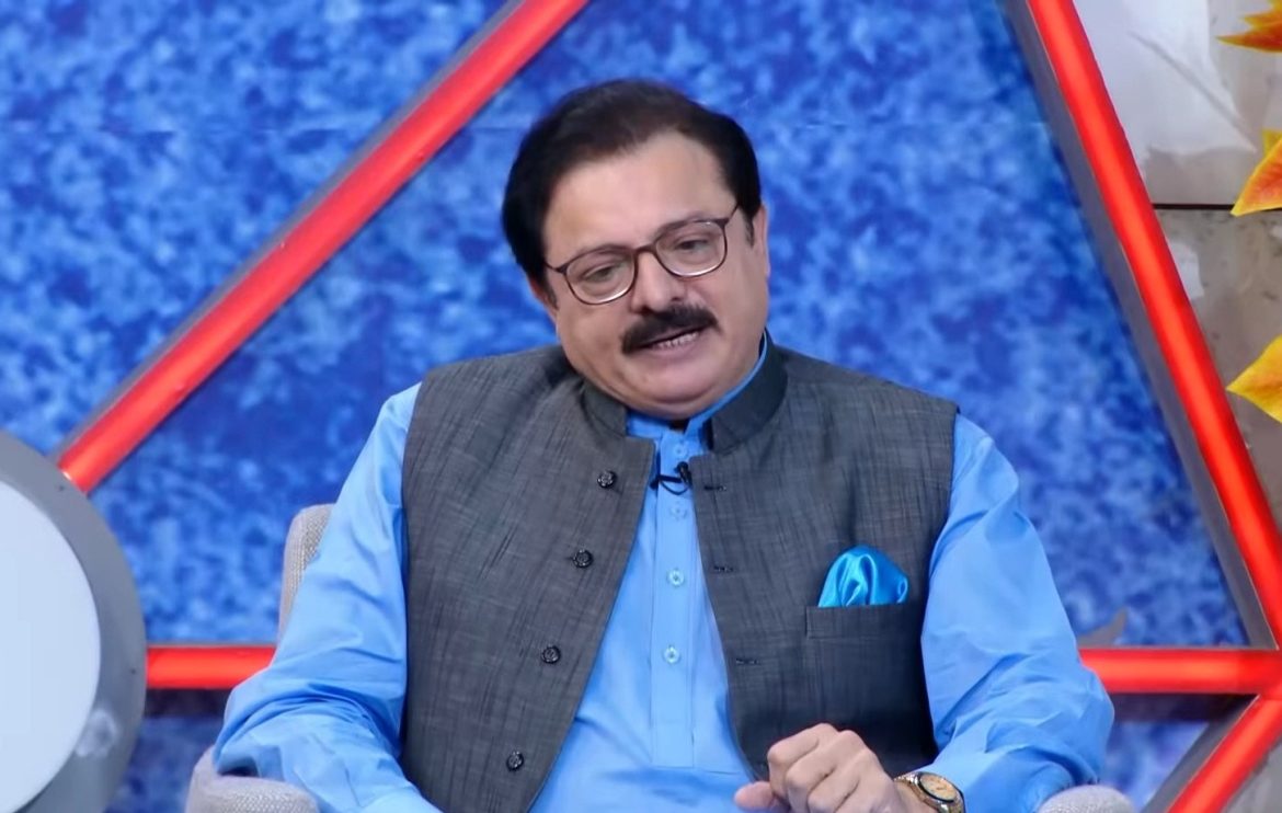 A person in a gray vest and light blue shirt sits on a set with a blue background and geometric designs, smiling and engaging.