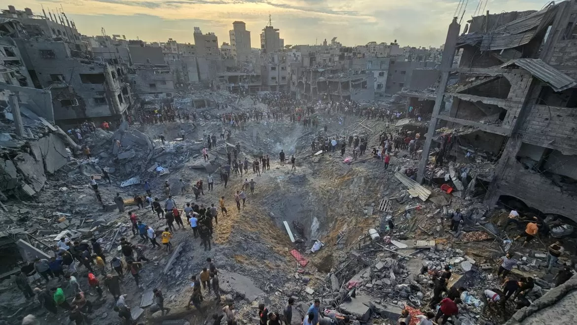 Crowds gather around a large bomb crater amidst the wreckage of destroyed buildings in a war-torn urban area at sunset.