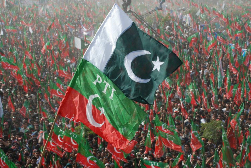 A large crowd waving green and red flags at a political rally, with prominent displays of the Pakistan flag and the PTI emblem.
