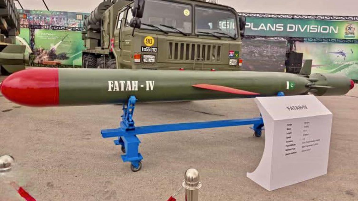 A Fatah-IV missile displayed on a blue stand, with specifications nearby, set against military vehicles and a promotional backdrop.