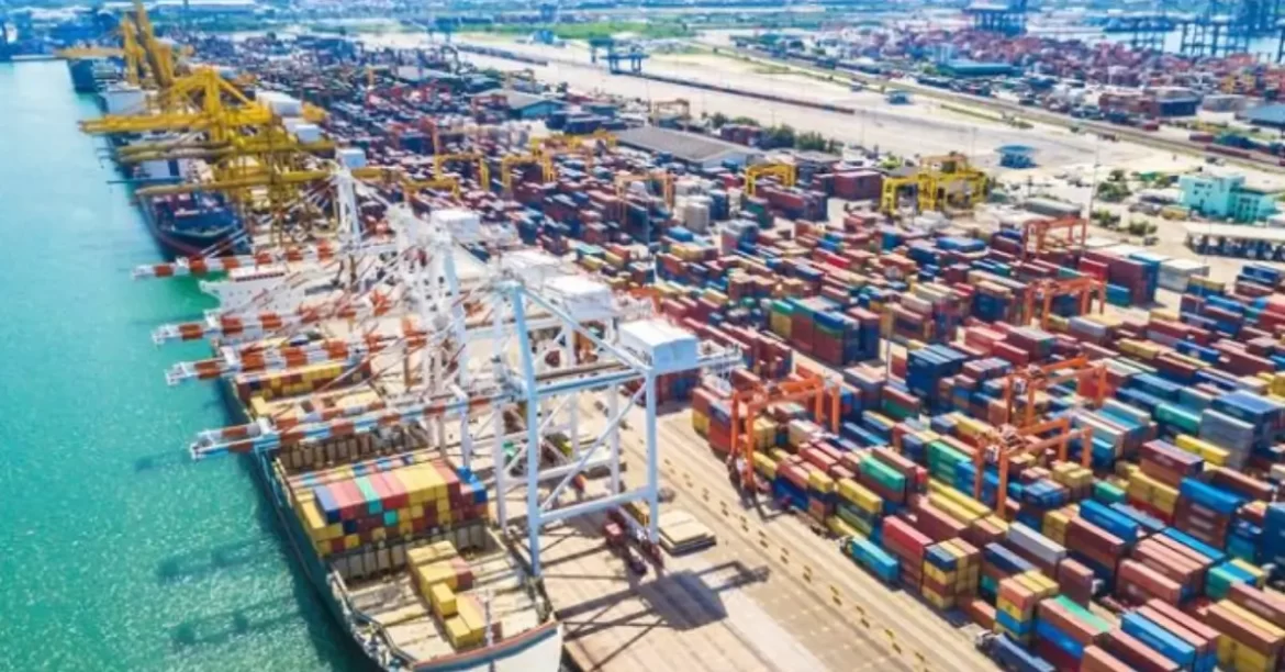 Aerial view of a bustling shipping port filled with colorful container stacks, cranes, and ships along a turquoise waterfront.