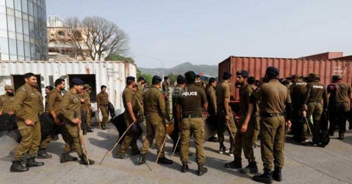 A group of police officers in brown uniforms gathers near shipping containers, equipped with batons, in an outdoor setting.
