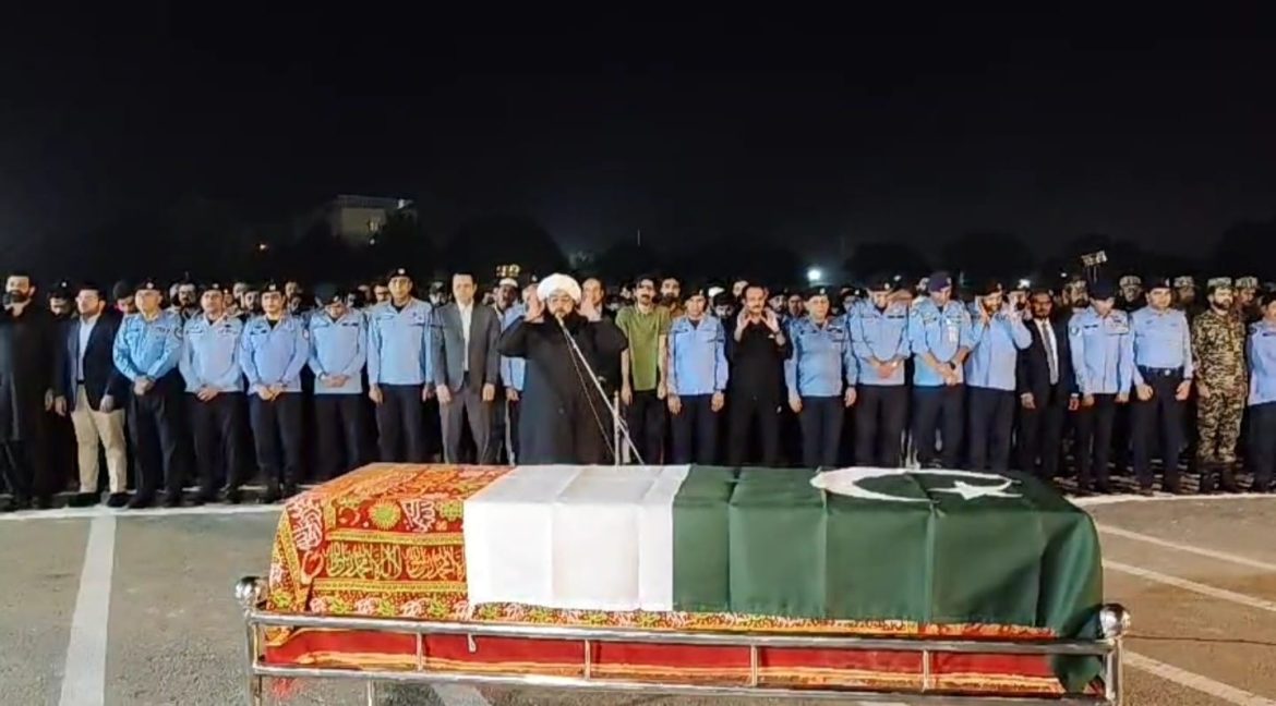 A solemn night scene with a covered coffin draped in a Pakistani flag, surrounded by a crowd in formal and police uniforms.