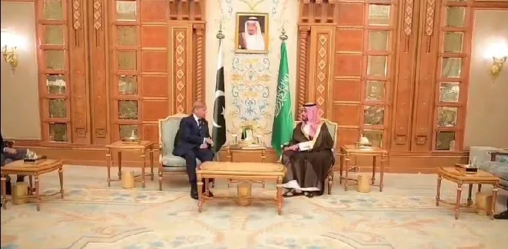 Pakistani and Saudi officials engage in discussion in an ornate meeting room, flags of both countries displayed prominently.