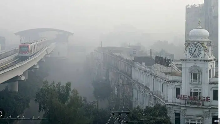 A train travels on an elevated track through a dense fog, revealing an old clock tower and buildings shrouded in smog.