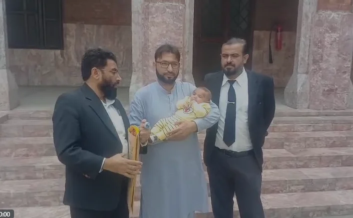 Three men in formal attire stand together on a stone staircase, one holding a wrapped baby, with documents in another man's hand.