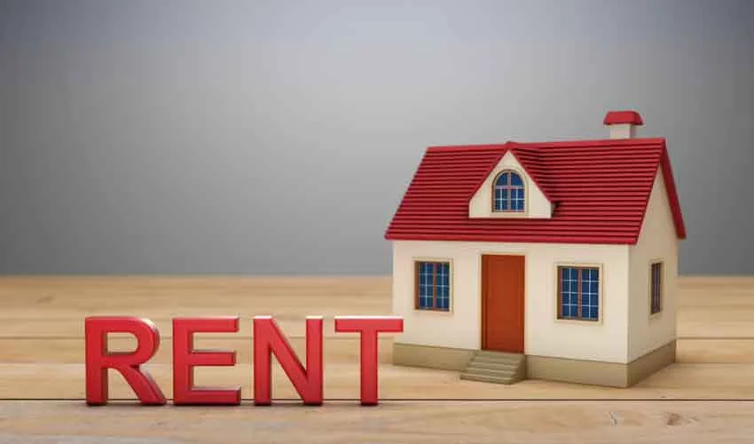 A small model house with a red roof sits next to the bold word RENT in bright red letters on a light wood surface.