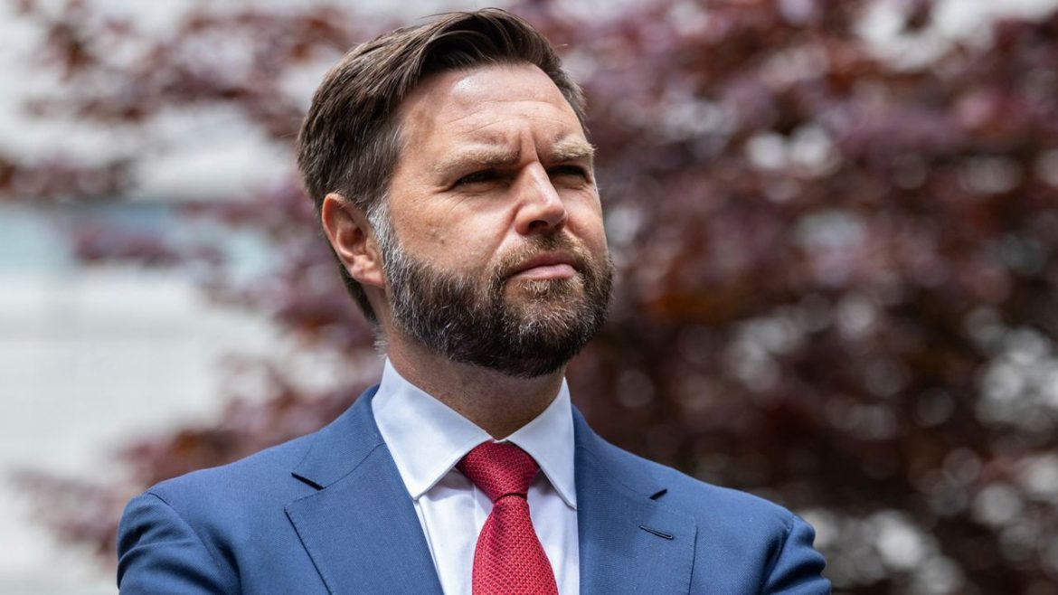 A well-dressed man in a suit and red tie stands with arms crossed, set against a blurred backdrop of red foliage.