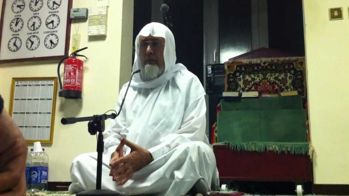 A person dressed in a white robe sits cross-legged in a room with a microphone, surrounded by clocks and shelves of books.