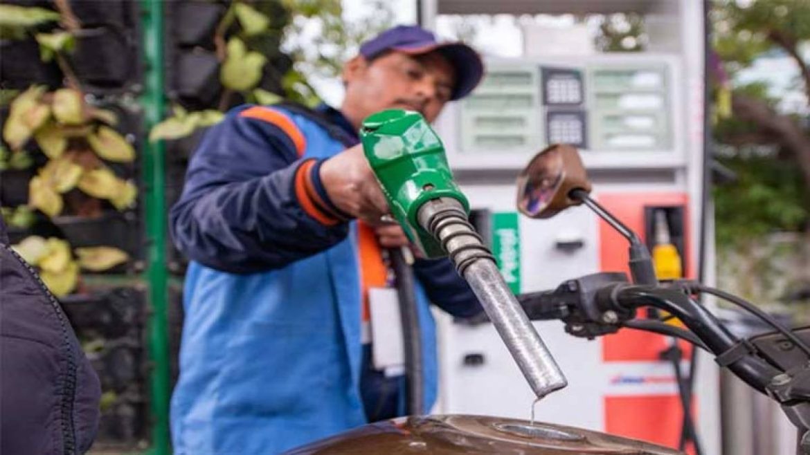A fuel attendant holds a green fuel nozzle above a motorcycle, ready to refuel at a gas station surrounded by greenery.