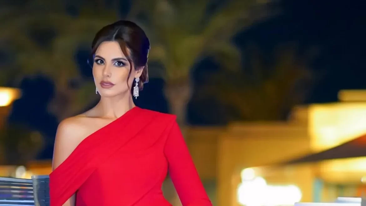 A woman in a red off-shoulder gown poses elegantly against a backdrop of palm trees and warm, ambient lighting.