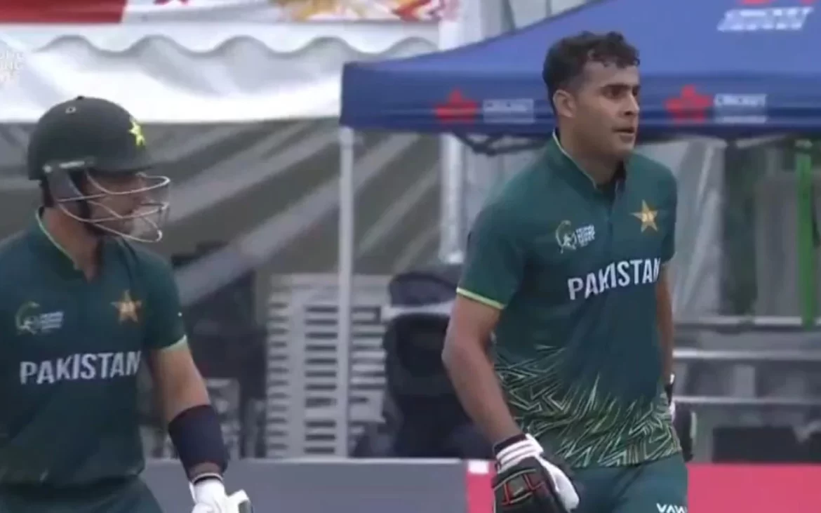 Two cricketers in green Pakistan jerseys walk together on a cricket field, with a tent and equipment in the background.