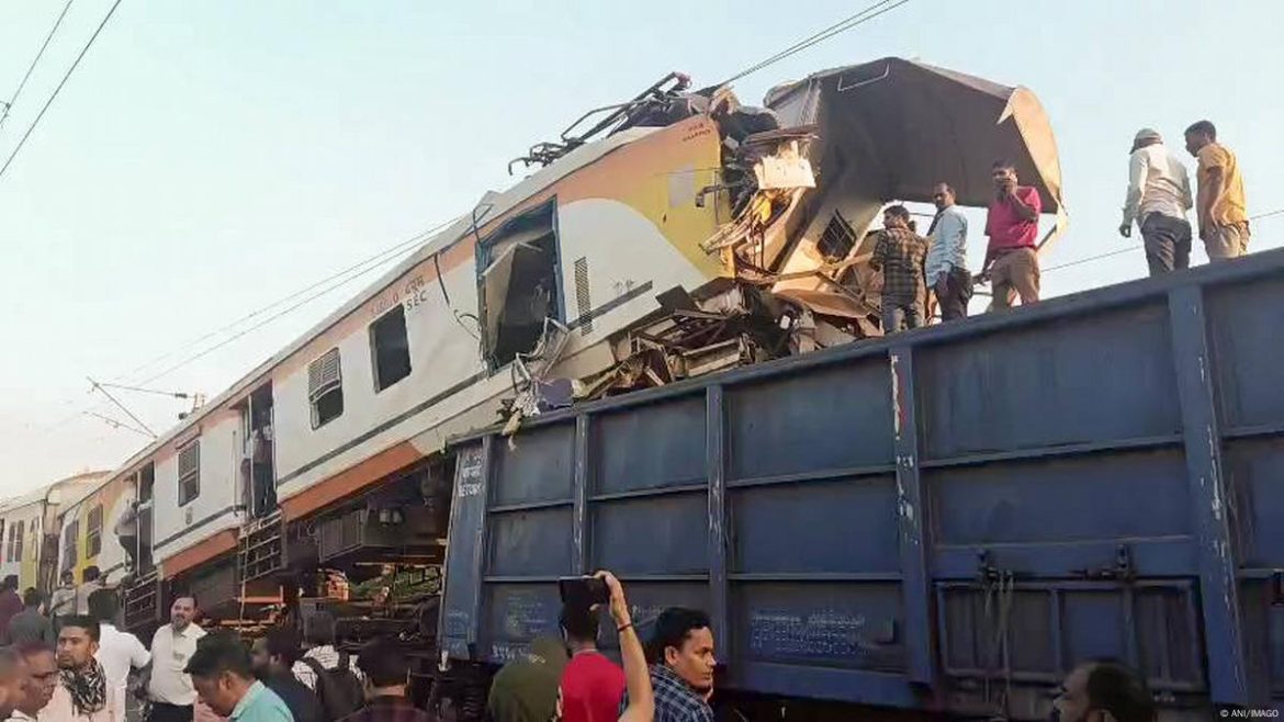A damaged train collides with a freight truck, with onlookers gathering around the scene amid a clear sky.
