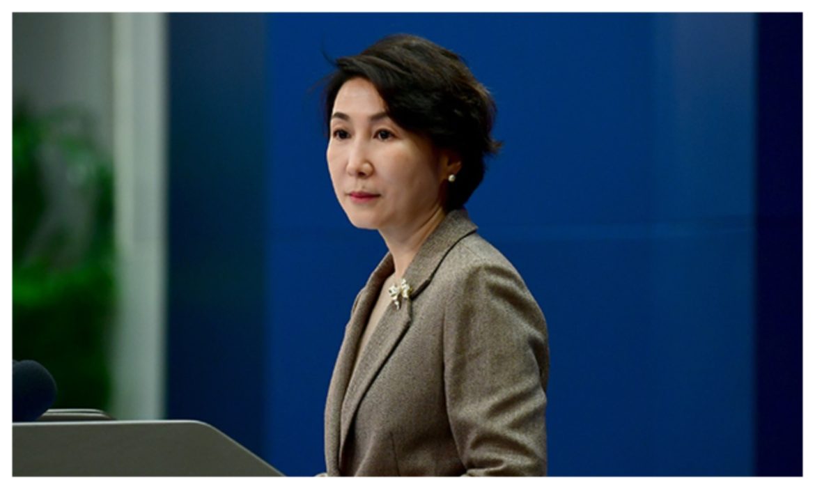 A woman in a gray blazer stands at a podium, addressing an audience with a blue backdrop and hints of greenery in the background.