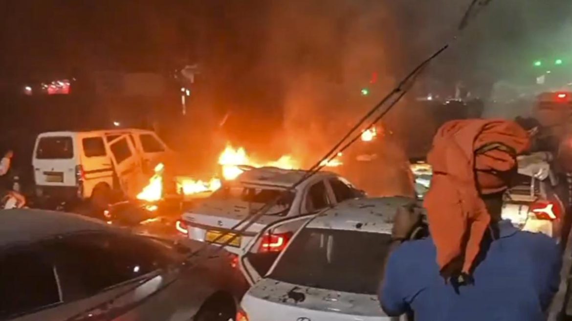 A chaotic scene of burning cars at night, with flames and smoke rising amidst a crowd of distressed onlookers.
