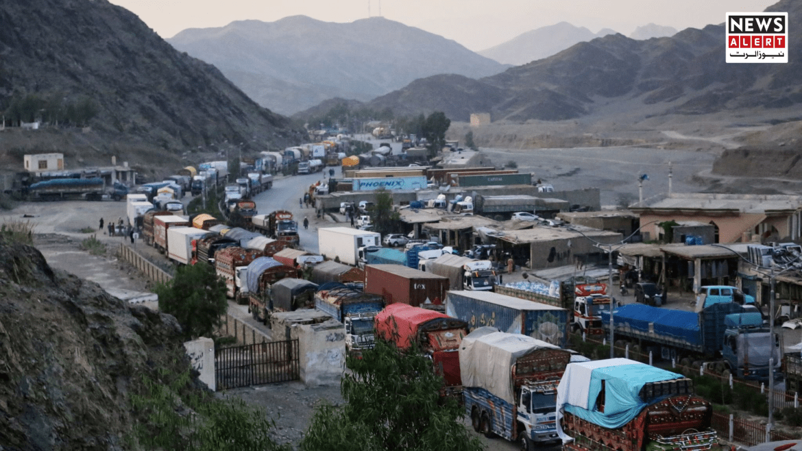 A long line of trucks and vehicles waits along a mountain road, with small shops and structures visible amidst the rugged terrain.
