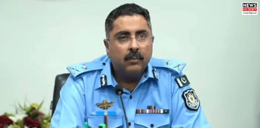 A police officer in a blue uniform sits at a conference table with flowers in the background, addressing an audience during a press briefing.