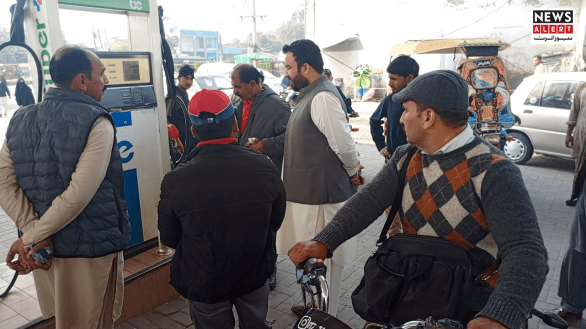 A group of men gathered near a fuel station, discussing something while one man holds a bicycle. Vehicles are parked in the background.