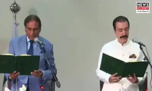 Two officials stand at microphones, holding green books during a formal ceremony, with a backdrop of flags and floral arrangements.