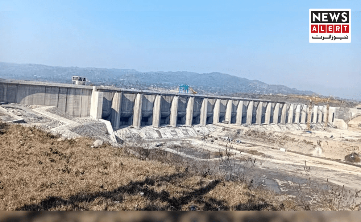 A large concrete dam against a blue sky, with a mountainous background and construction activity visible on top.