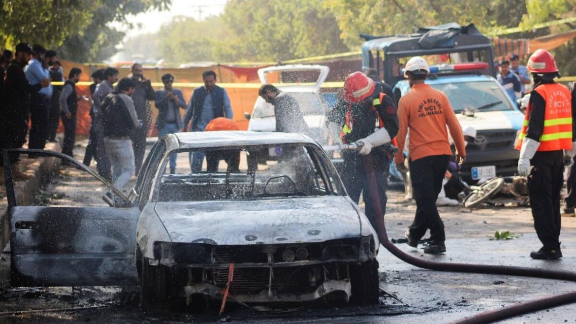 Emergency responders douse a burned-out car with water while a crowd watches nearby at the scene of an incident.