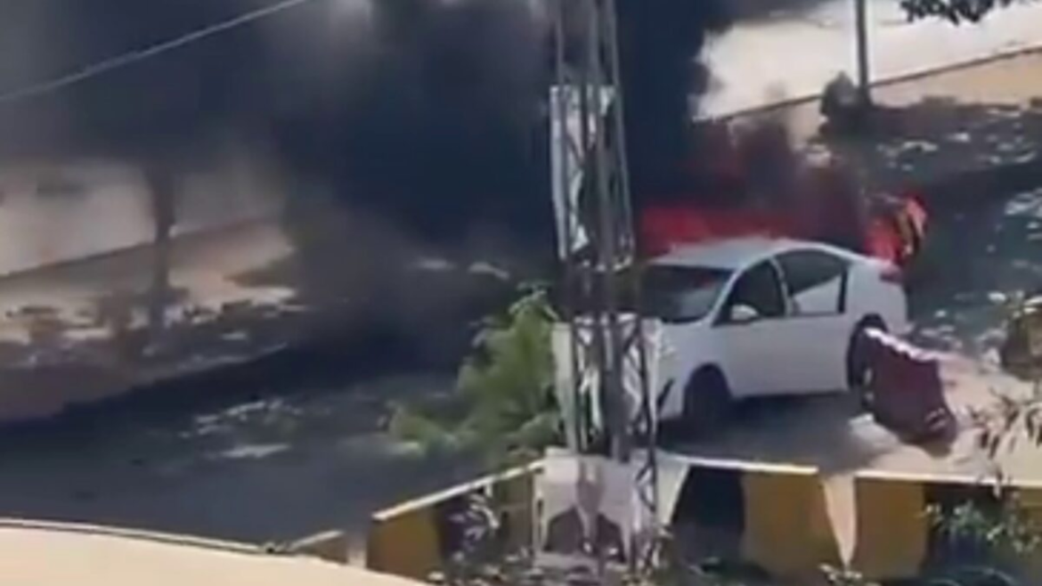 A white car sits on the street, partially obscured by thick black smoke and flames in the background, indicating a fire or explosion.