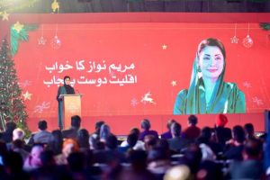 A speaker stands at a podium in front of a festive backdrop, addressing an audience with a large screen displaying text in Arabic.