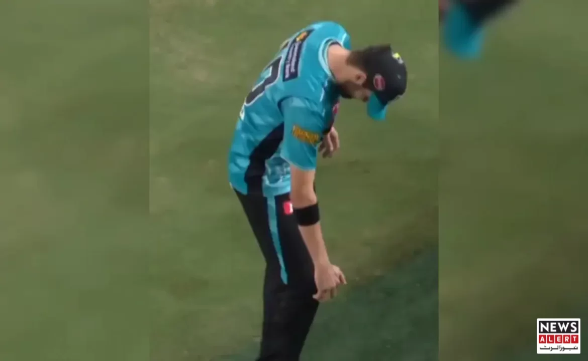 A cricket player in a blue uniform leans down, inspecting his leg on a grassy field during a match.