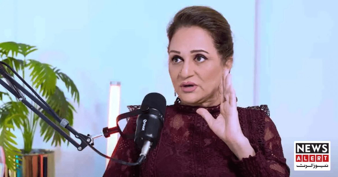 A woman in a lace burgundy top speaks into a microphone, gesturing with her hand, surrounded by greenery and modern decor.