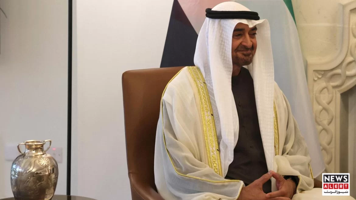 A man in traditional attire sits in a chair, with a decorative silver jug in the foreground and flags in the background.