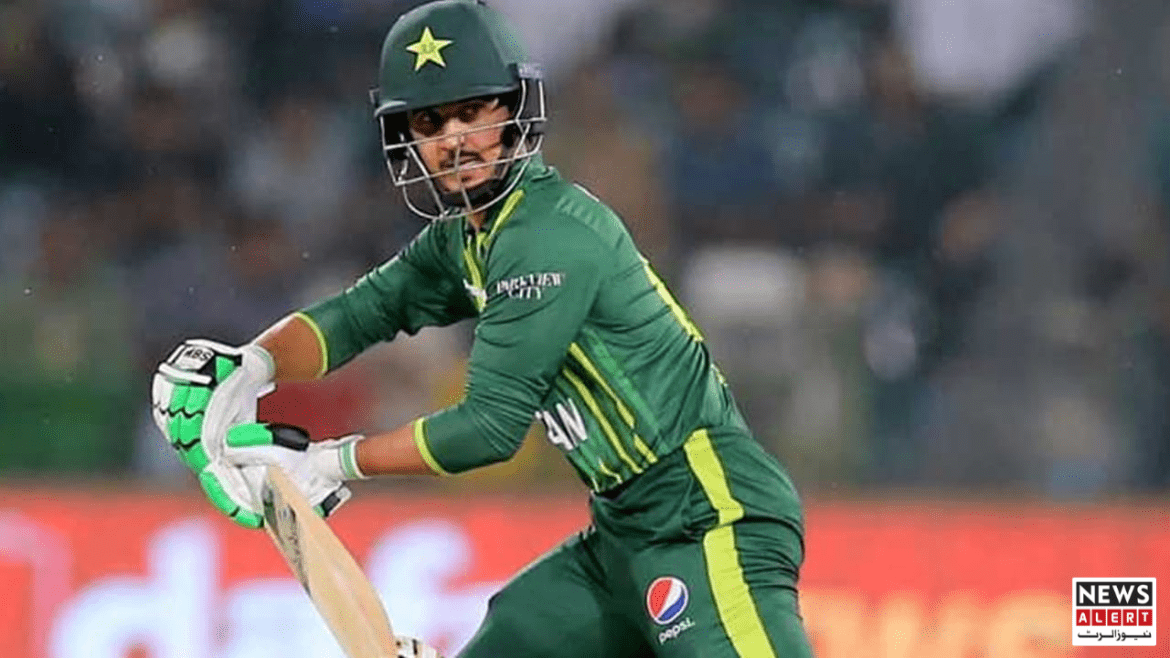 A cricketer in a green uniform prepares to hit the ball, with a blurred crowd in the background.