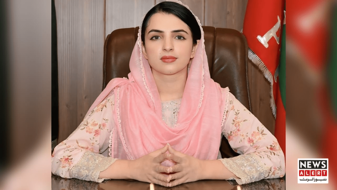 A woman in a floral dress and pink shawl sits at a wooden desk, hands clasped, with flags in the background.