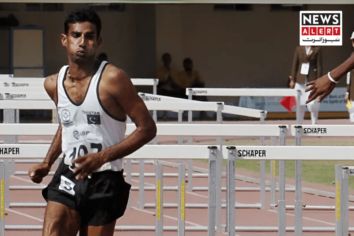 A male athlete in a tank top runs towards a series of hurdles on a track, with officials and spectators visible in the background.