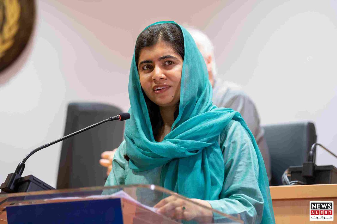 A woman in a turquoise shawl speaks at a podium, with equipment in the background and a blurred figure seated behind her.